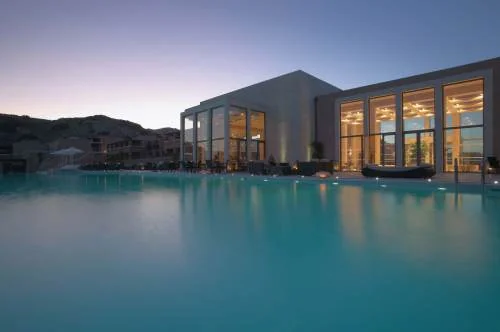 Illuminated glass-fronted building beside a swimming pool at dusk at Helona Resort, Kos.