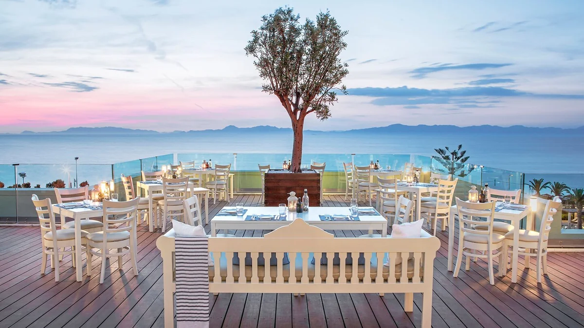 White tables and chairs on a restaurant terrace overlooking the sea in Rhodes at sunset.